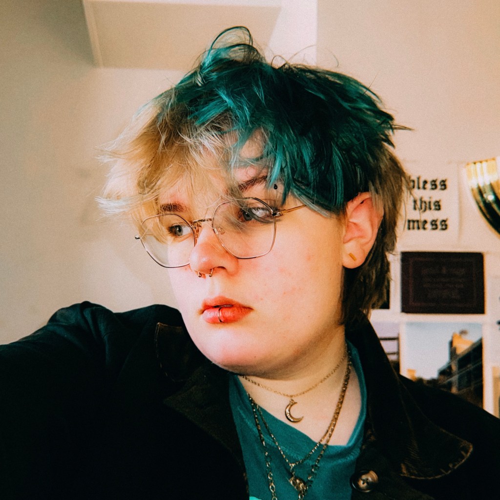 A photo of Amara Rosenau looking to the left of the camera. Amara has a green shirt, black coat, layered gold necklaces, and gold glasses.